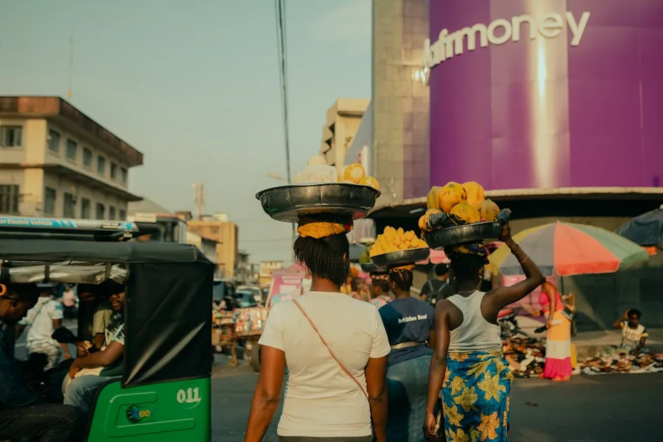 Women carrying food on their heads walk through a market.